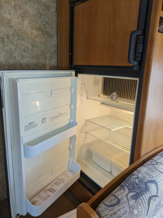Inside an rv rental: an open, empty Dometic RV refrigerator with a thermometer, featuring wood-look doors and wire shelves.. Coachmen Freelander Deluxe/Premier 2005