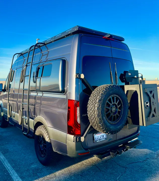 This custom dark gray camper van, an ideal RV rental option, features a roof rack, ladder, and rear-mounted spare tire for adventure.. Mercedes-Benz Sprinter 2021