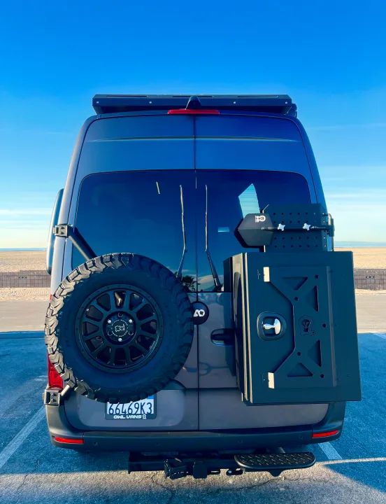 Rear of a gray RV rental van with a spare tire and storage box. Parked at a sunny beach, ready for adventure.. Mercedes-Benz Sprinter 2021
