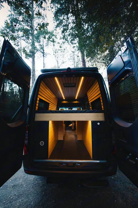 Spacious converted RV rental van interior, viewed from the rear, with warm lighting, bed, and ample storage. Parked in nature.. Mercedes-Benz Sprinter 2021