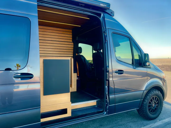 Spacious RV rental van with a sleek blue exterior, modern wood interior, and off-road tires, parked by a sunny beach.. Mercedes-Benz Sprinter 2021