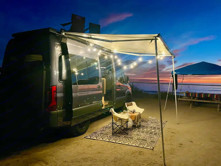 An RV rental with an illuminated awning creates a cozy evening scene by the ocean. String lights glow over a rug and chairs.. Mercedes-Benz Sprinter 2021