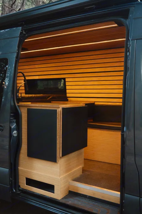 Modern rv rental camper van interior: light wood walls, LED lighting, compact kitchen with black cabinets, and bench.. Mercedes-Benz Sprinter 2021