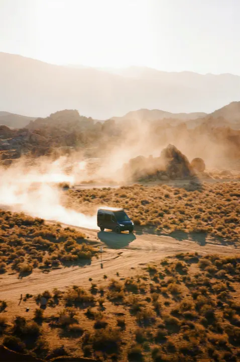 An RV rental van kicks up dust on a remote desert road, surrounded by rugged hills and warm, hazy light. Off-road travel.. Mercedes-Benz Sprinter 2021