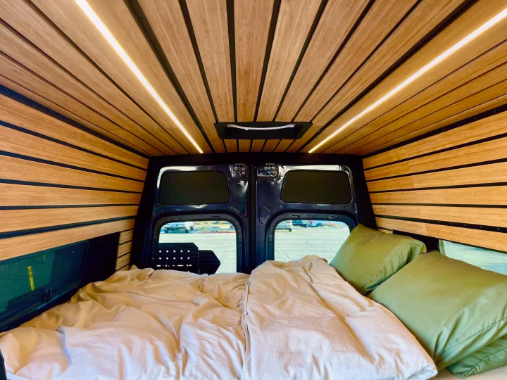 Modern RV rental bedroom featuring sleek wood paneling, ambient LED lighting, and a cozy bed with green pillows.. Mercedes-Benz Sprinter 2021