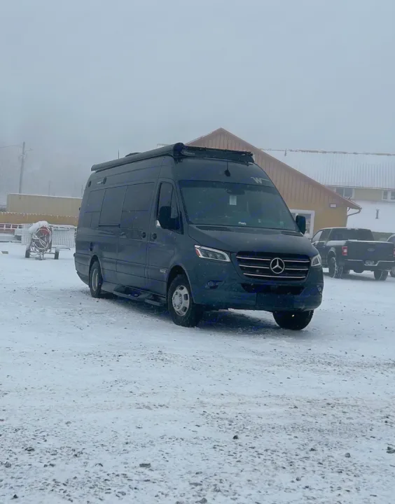 Dark Mercedes-Benz Sprinter rv rental camper van parked on snow in a winter setting. Buildings and a pickup are in the background.. Winnebago Era 2021
