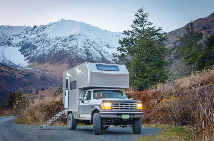 This custom truck camper RV rental is ready for mountain adventure. Parked on a gravel road, headlights on, with snowy peaks.. Ford F250 1997