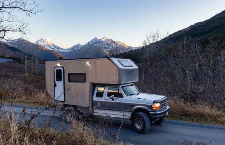 This custom truck camper, an RV rental, rests on a mountain road with snow-capped peaks behind.. Ford F250 1997