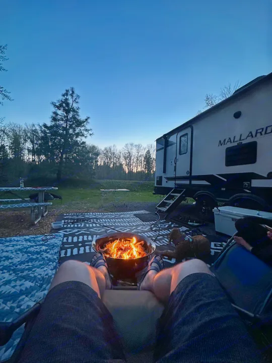 Serene dusk at an RV rental campsite. View from person's perspective, campfire warming legs, Mallard RV nearby.. Heartland RVs Mallard 2021