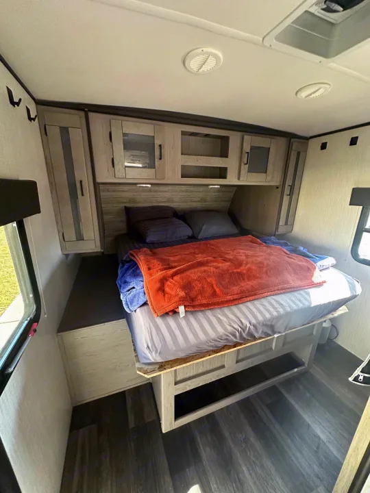 RV rental bedroom: striped bed with orange blanket, dark pillows, wood-look floor, and light wood overhead cabinets.. Heartland RVs Mallard 2021