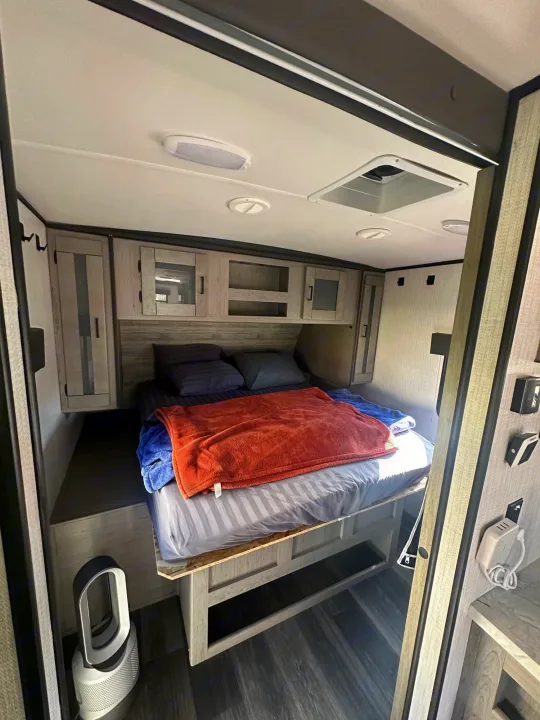 Cozy RV rental bedroom featuring an elevated bed with storage, overhead cabinets, and a Dyson fan. Ideal for compact living.. Heartland RVs Mallard 2021