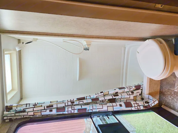 Inside this RV rental, a compact bathroom reveals a white tiled shower, toilet, and patterned shower curtain.. Coachmen Apex 50th Anniversary 2015