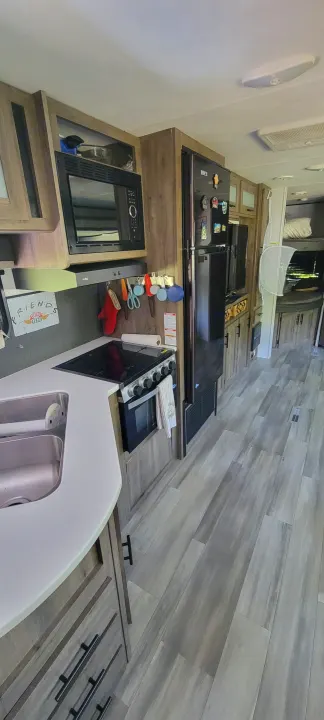 Modern RV rental interior: kitchen with a full stove, microwave, fridge, and sink. Wood-look floors lead to bunk beds.. Heartland RVs Pioneer 2021
