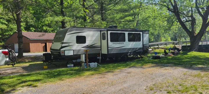 An RV rental (Pioneer travel trailer) at a sunny, treed campground with a truck, bike, and outdoor setup for relaxation.. Heartland RVs Pioneer 2021