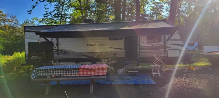 Sunny RV rental campsite setup with picnic table, grill, and blue mat. Pioneer travel trailer nestled in nature.. Heartland RVs Pioneer 2021