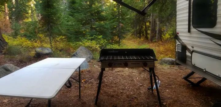 RV rental camping setup: a white folding table, Camp Chef outdoor stove, and travel trailer nestled in a beautiful forest.. Jayco Jay Flight SLX 2019