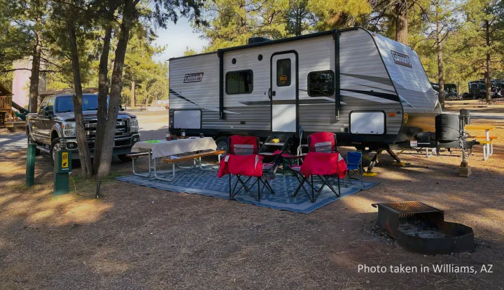 Relaxing RV rental campsite in Williams, AZ with a travel trailer, truck, picnic area, and fire pit under pine trees.. Dutchmen Coleman Lantern LT 2021