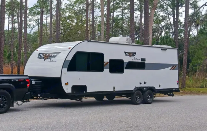 Considering an RV rental? This white travel trailer, towed by a black truck, is ready for your next camping trip in the forest.. Forest River Wildwood X-Lite Platinum 2024