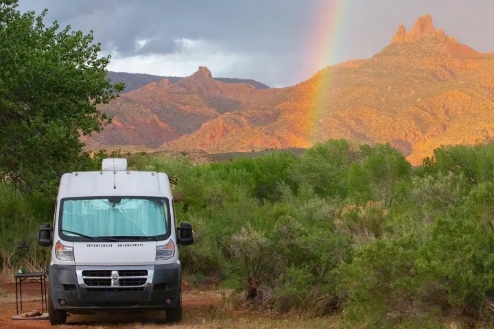 Scenic view of an RV rental nestled among green bushes, featuring a colorful rainbow arching over rugged red mountains.. Dodge Sprinter Van 2016