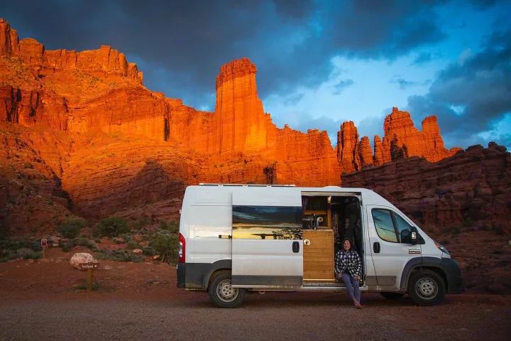 Red rock van life: An RV rental parked at sunset, a woman sits in its open door admiring the stunning desert scenery.. Dodge Sprinter Van 2016
