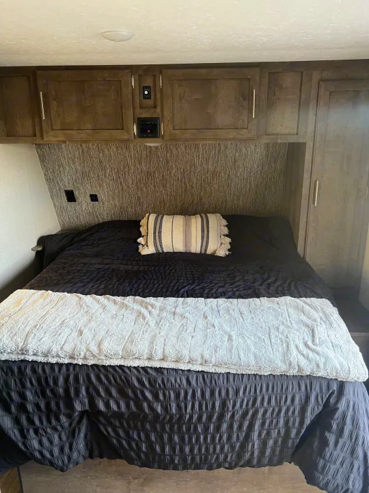Cozy RV rental bedroom featuring a bed with dark bedding, striped pillow, and wood overhead cabinets for storage.. KZ Escape 2019