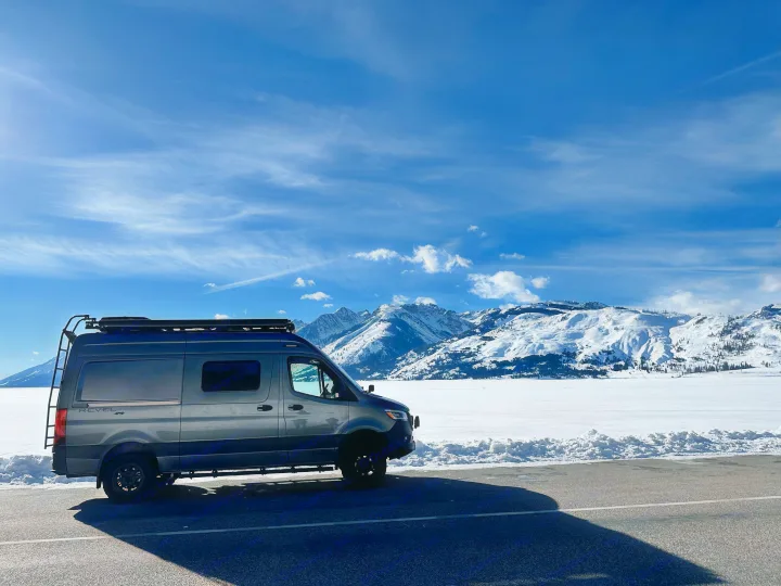 Discover winter with an rv rental! A gray camper van sits on asphalt beside a frozen lake & snow-covered mountains.. Winnebago Revel 2024