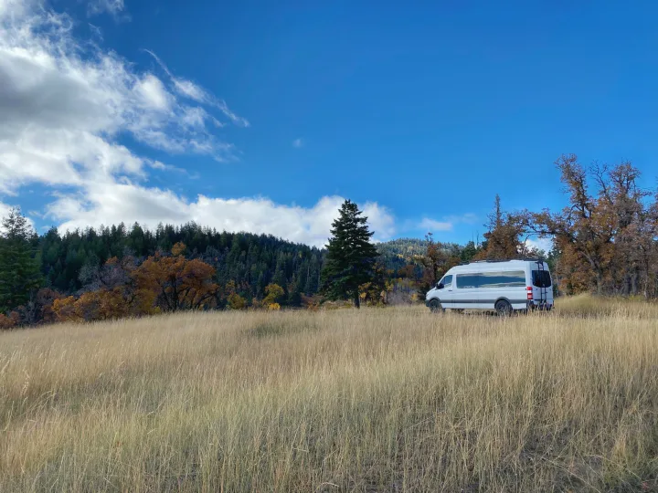 White RV rental parked in a tall grassy field with autumn trees under a blue sky, perfect for nature travel.. Mercedes-Benz Sprinter 2014