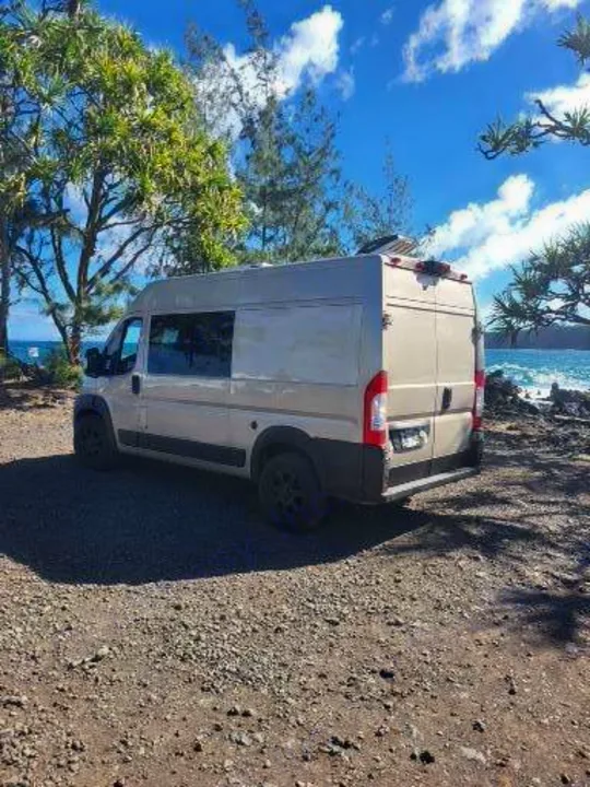 This RV rental, a beige camper van, is parked oceanfront, surrounded by lush trees under a bright blue sky.. Dodge Ram Promaster 2021