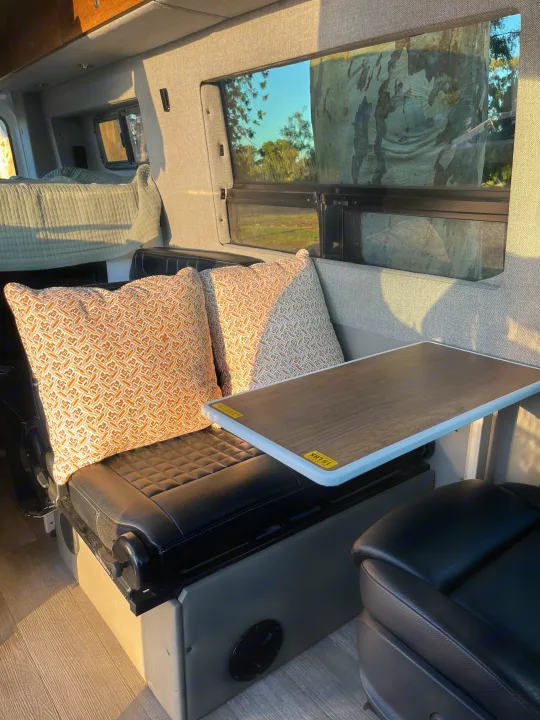 Cozy RV rental interior showing a bed, patterned pillows, table, and seating by a window with tree views.. Storyteller Overland Storyteller Overland Class B 2020