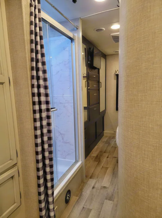 This RV rental interior features a modern shower with a checkered curtain, a compact hallway, and built-in storage leading to the bedroom.. Jayco Greyhawk 2022
