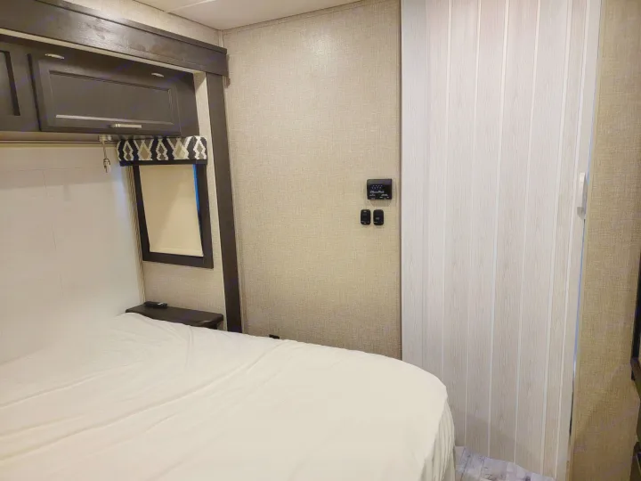Cozy RV rental bedroom featuring a clean bed, dark overhead cabinets, and wall-mounted controls. Ready for your journey!. Jayco Greyhawk 2022