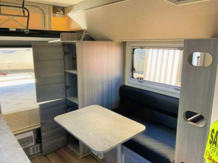 Inside this RV rental: a compact dinette with a table and dark seating, open cabinets, and a bed. Wood-look floor.. OBI OBI Popup Trailer 2022