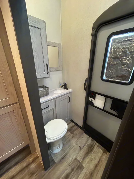 Clean and compact RV rental bathroom with a toilet, sink vanity, mirror, and modern gray wood-look flooring.. Keystone RV Montana High Country 2023