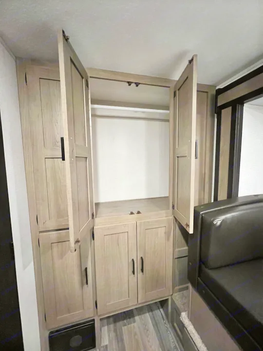 RV rental interior features light wood cabinetry: An open wardrobe with a hanging rod, lower storage, and a black cushioned seat.. Keystone RV Hideout 2021