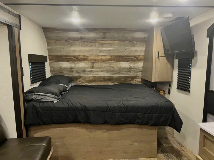 Cozy RV rental bedroom featuring a bed with black bedding, wood accent wall, mounted TV, and small windows.. Keystone RV Hideout 2021