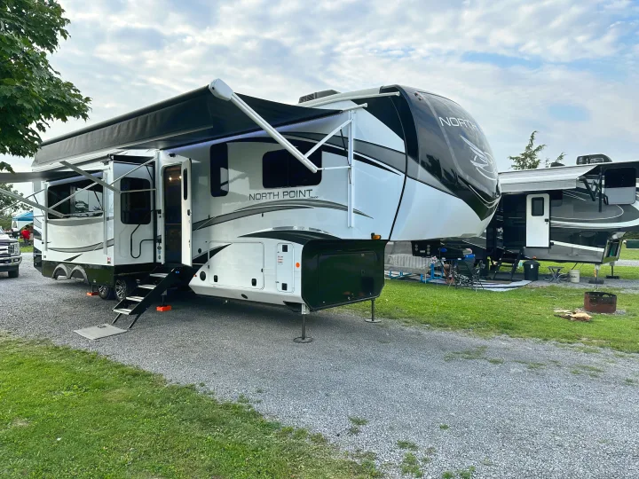 A large, modern white and black fifth-wheel RV is set up at a gravel campsite, perfect for an rv rental getaway.. Jayco North Point 2023