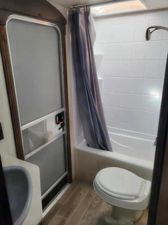 An RV rental's compact bathroom features a white shower/tub combo with a gray curtain, a white toilet, and a small sink.. Forest River Cherokee Grey Wolf 2019