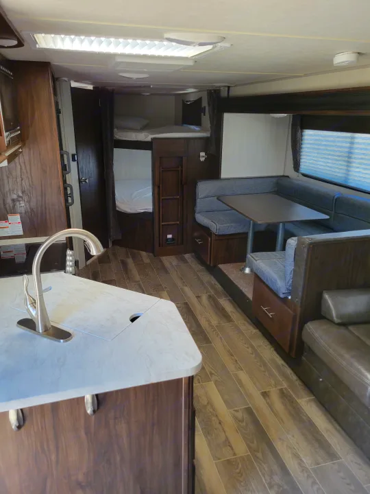 Inside an RV rental, showing the kitchen counter, dining area with table & benches, and bunk beds. Wood-look flooring and bright ceiling lights.. Forest River Cherokee Grey Wolf 2019