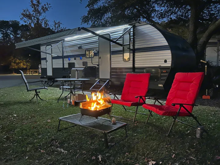An RV rental setup at dusk: an illuminated camper with awning, a warm campfire, and red chairs on grass. Great for camping.. Forest River Cherokee Grey Wolf 2019