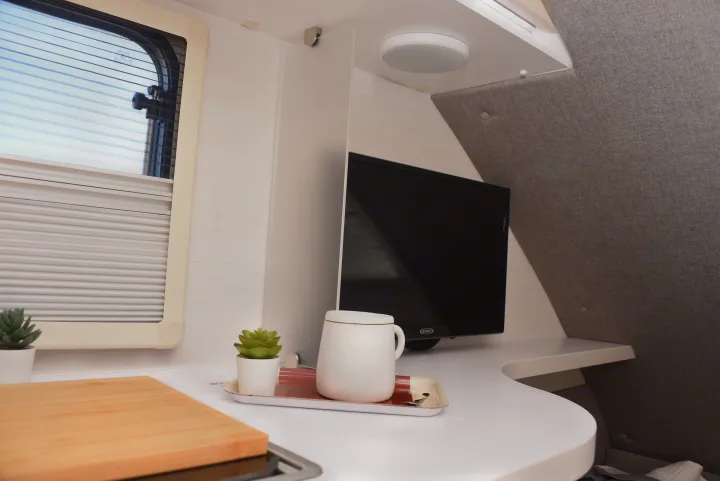 Bright rv rental interior showing a window, flat-screen TV, and a clean white counter with a decorative plant and mug.. Nucamp Tab 400 2024