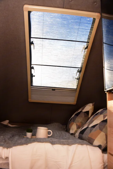 Cozy RV rental interior featuring a made bed with patterned pillows, a window with blinds, and a mug with a plant.. Nucamp Tab 400 2024