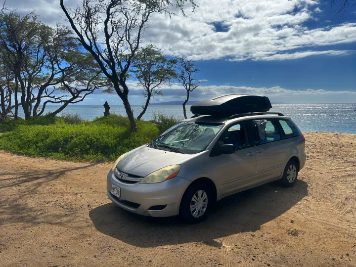 Silver minivan with roof cargo box parked on a dirt road by the ocean; like an RV rental for beach adventures.. Toyota Other 2006