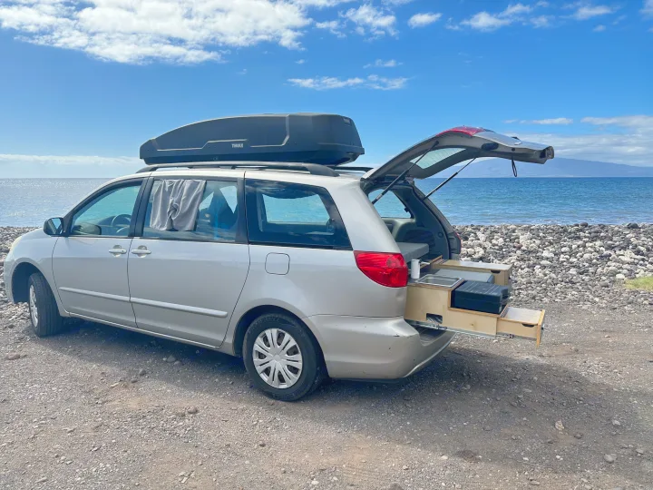 An RV rental: a converted minivan with roof box & pull-out kitchen, parked by a rocky beach.. Toyota Other 2006