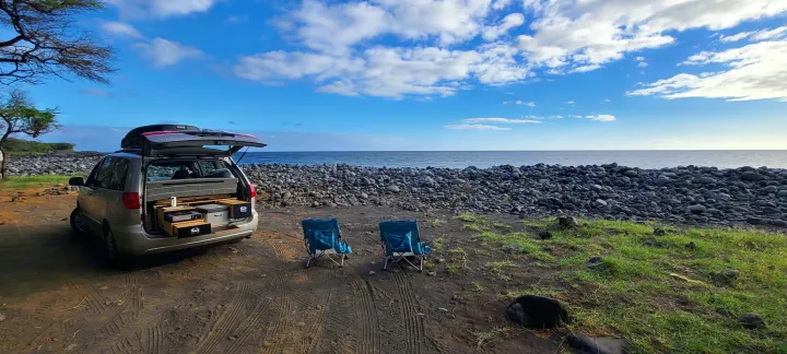 Custom minivan setup with an open tailgate kitchen, and two chairs facing a rocky beach and ocean. A versatile RV rental alternative for coastal trips.. Toyota Other 2006