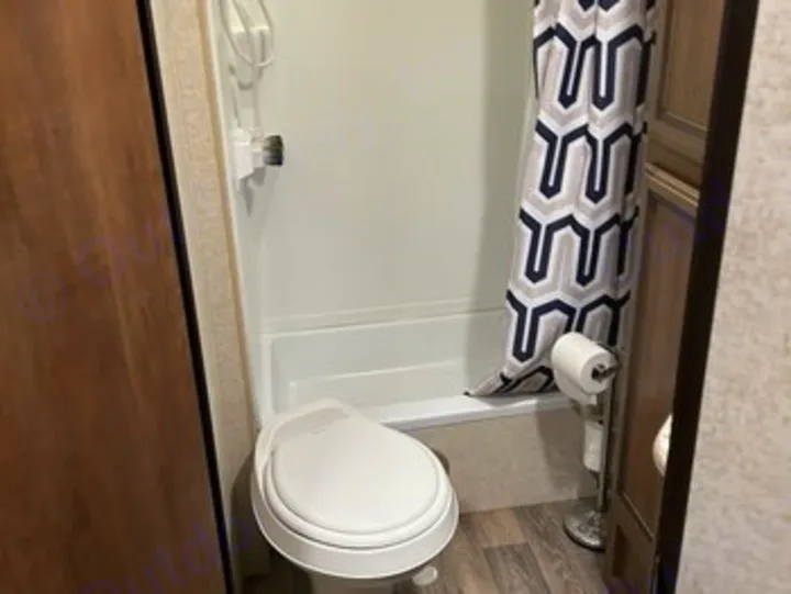 A compact RV rental bathroom with a toilet and small shower/tub combo, featuring a geometric shower curtain and wood-look flooring.. Heartland RVs Sundance XLT 2016