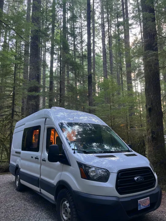 White rv rental van in a tall BC forest, interior lights on. Great for forest camping and adventure.. Ford Transit 2019