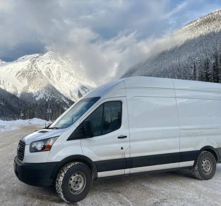 A white RV rental van is parked on a snowy road, surrounded by majestic snow-capped mountains and a winter forest.. Ford Transit 2019