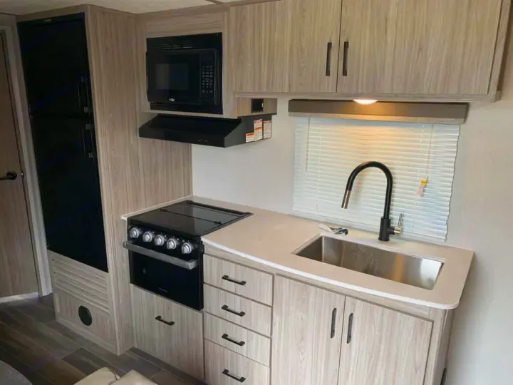 Explore this modern rv rental kitchen: fridge, microwave, oven, and sink under a window with blinds. Compact and functional.. Keystone RV Passport SL 2021