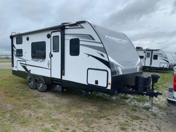 White & gray Passport travel trailer RV rental, featuring an awning & dual axles, parked on a grassy lot.. Keystone RV Passport SL 2021