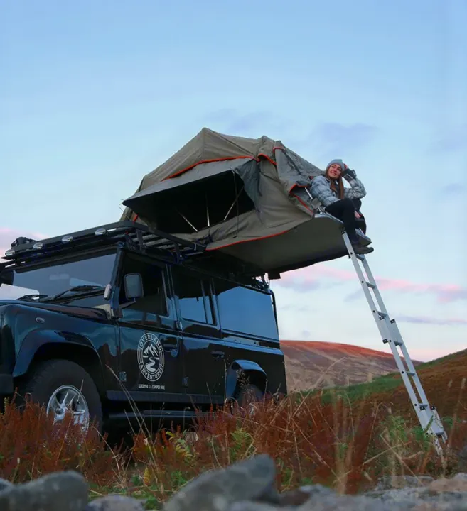 Experience overland camping with our luxury rv rental! A woman relaxes on a rooftop tent mounted on a black 4x4 camper hire SUV.. Land Rover 2013 Land Rover Defender 110 XS Utility + 1 Roof Tent 2016